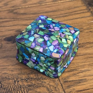 May include: A square jewelry box with a mosaic design. The box is covered in small, irregularly shaped pieces in shades of blue, green, and purple. The box sits on a wooden surface.
