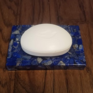 May include: A white oval bar of soap rests on a rectangular soap dish. The soap dish is made of blue and gray stones set in a resin base. The soap dish is on a wooden surface.