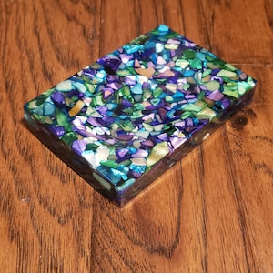 May include: A rectangular soap dish with a mosaic design. The dish is filled with small, irregularly shaped pieces in shades of blue, green, purple, and white. The dish is set on a wooden surface.