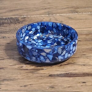 May include: A small, round bowl with a mosaic design. The bowl is primarily blue with accents of white and gray, creating a textured appearance. The bowl sits on a wooden surface.