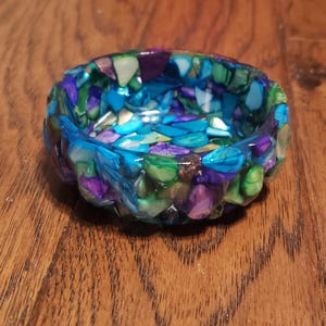 May include: A small, round bowl made of resin with a mosaic design. The bowl is filled with small, colorful pieces of glass and has a blue, green, and purple color scheme.