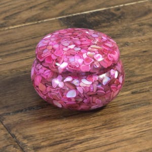 May include: A small, round, pink decorative box with a lid. The box is covered in small, irregular pieces of pink and white material, creating a mosaic-like pattern. The box sits on a wooden surface.