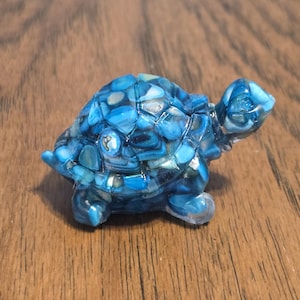 May include: A small, decorative turtle figurine. The turtle's shell is a mosaic of blue and turquoise stones, with a clear, glossy finish. The head, legs, and tail are also clear, giving the figurine a three-dimensional appearance. The figurine is on a wooden surface.
