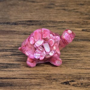 May include: A small, decorative turtle figurine. The turtle is primarily pink with a shell composed of smaller, lighter pink and white pieces. The figurine has a glossy finish and is set against a wooden background.