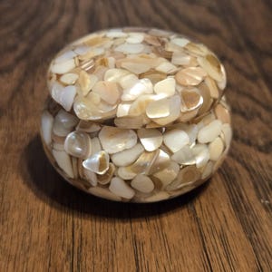 May include: A small, round trinket box made of iridescent white and beige shell pieces. The box has a lid and is perfect for storing small items.
