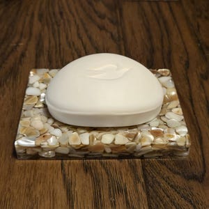 May include: A white soap bar with a dove logo sits on a square soap dish made of iridescent seashells.