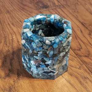 May include: A hexagonal pen holder made of clear resin embedded with blue, gray, and white pebbles. The container has a smooth, polished finish and sits on a wooden surface.