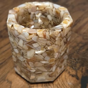 May include: A hexagonal shaped container made of clear resin with embedded seashells. The container is filled with small, white and beige seashells.