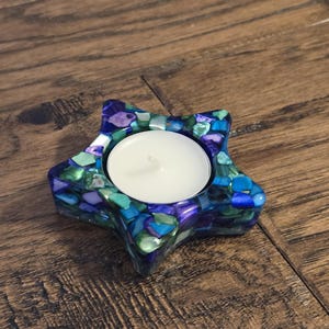 May include: A star-shaped tealight candle holder with a mosaic design. The holder is filled with blue, green, and purple stones. A white candle sits in the center of the holder. The holder is on a wooden surface.