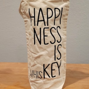 May include: A beige canvas wine bottle bag with the words "HAPPINESS IS KEY WHISKEY" printed in black. The bag is standing upright on a wooden surface.