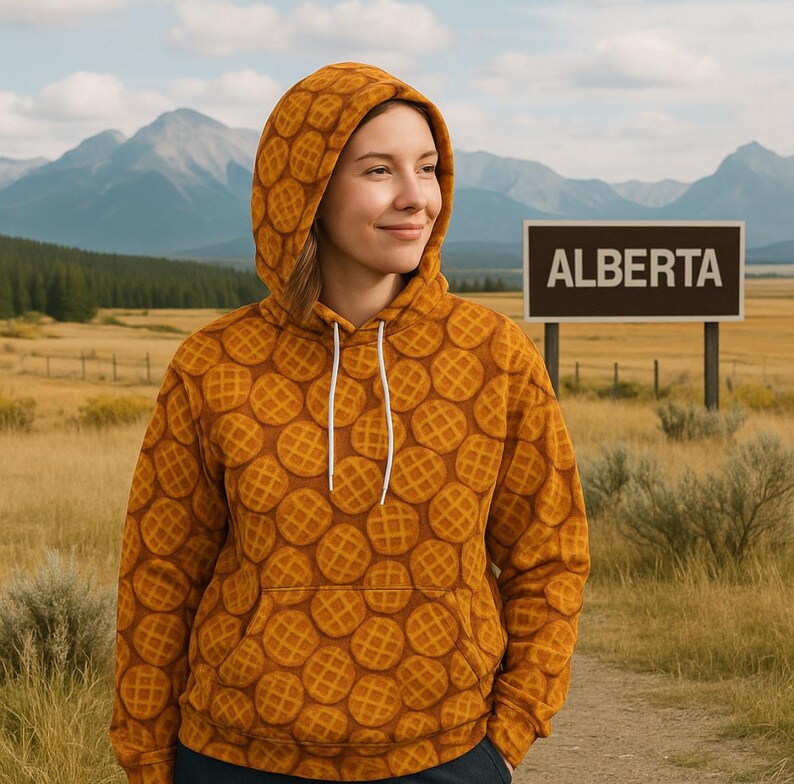 May include: A mustard-colored hoodie with a waffle pattern. The hoodie has a drawstring hood and a front pocket. The word "ALBERTA" is displayed on a brown sign in the background. The background includes mountains and a field.