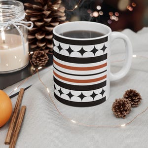 Puede incluir: Una taza de cerámica blanca con un patrón geométrico repetitivo de semicírculos y diamantes negros, acentuado por dos franjas horizontales de color naranja quemado. La taza tiene un asa estándar y está llena de un líquido oscuro.