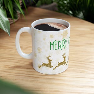 Puede incluir: Taza de cerámica blanca llena de café oscuro. La taza presenta el texto verde "MERRY" y diseños dorados de renos y copos de nieve. La taza tiene un asa blanca y está colocada sobre una superficie de madera.