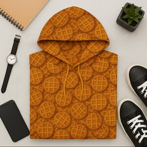May include: A hooded sweatshirt with a repeating pattern of round waffles in shades of brown and orange. The drawstring and hood lining match the waffle design. The sweatshirt is displayed with a watch, phone, sneakers, and a small plant.