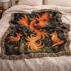 May include: Woven throw blanket with a phoenix design in orange and gold on a dark green backdrop. The blanket features a deer, fox, and forest details. It has a fringed trim around the edges. Measures approximately 152 cm x 127 cm.