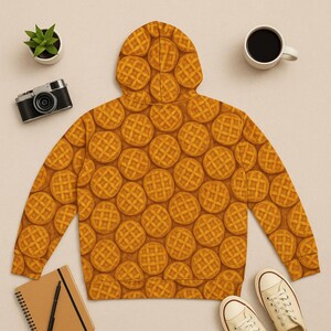 May include: A hooded sweatshirt featuring a repeating pattern of golden-brown waffles. The waffle design covers the entire garment, including the hood and sleeves. The sweatshirt is displayed on a neutral surface with a camera, coffee cup, plant, notebook, and shoes.