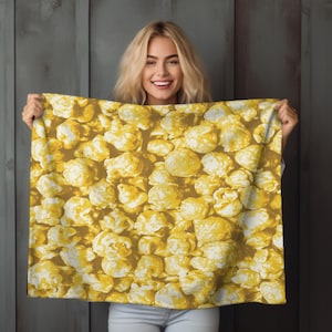 May include: A yellow and white popcorn-patterned blanket. The blanket features a repeating pattern of popcorn kernels in shades of yellow and white. The blanket is held up by a person.