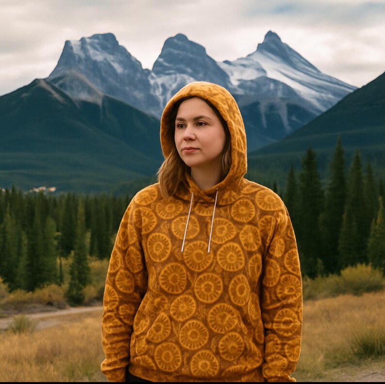 May include: Mustard-colored hoodie with a repeating circular pattern. The hoodie has a drawstring and a hood. The background features a mountain range and a forest. The hoodie is made of a soft, comfortable material.