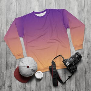 May include: A crew neck sweatshirt with a gradient design, transitioning from purple to orange. A gray and maroon baseball cap, a white disposable cup, and a black camera with a strap are also visible.