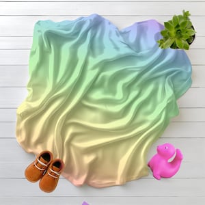 Op de afbeelding: Een bovenaanzicht van een vloeiende, iriserende stof in pastelkleuren geel, groen en blauw. Bruine leren babyschoenen met witte veters en een roze speelgoed dinosaurus liggen op een wit houten oppervlak, samen met een potplant.