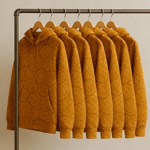 May include: Seven mustard-colored hoodies with a repeating circular pattern hang on wooden hangers from a metal clothing rack. Each hoodie has a front pocket and a hood. The hoodies are made of a soft material.