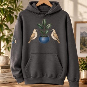 Peut inclure: Un sweat à capuche gris anthracite orné d'un motif central représentant deux oiseaux brun clair face à une plante verte dans un pot bleu. Une version plus petite du même motif d'oiseau et de plante est imprimée sur la manche droite près du poignet.