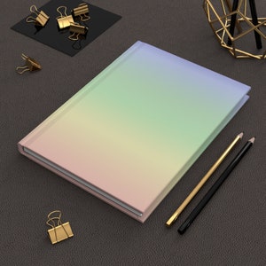 May include: A hardcover journal with a pastel rainbow gradient cover. Gold and black pencils and gold binder clips are arranged around the journal on a dark gray surface. The journal is closed.