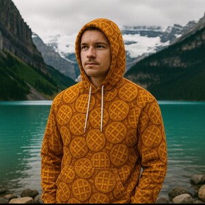 May include: A mustard-colored hoodie with a waffle pattern. The hoodie has a drawstring hood and a front pocket. The waffle design is a repeating pattern of circular waffles. The background features a lake and mountains.