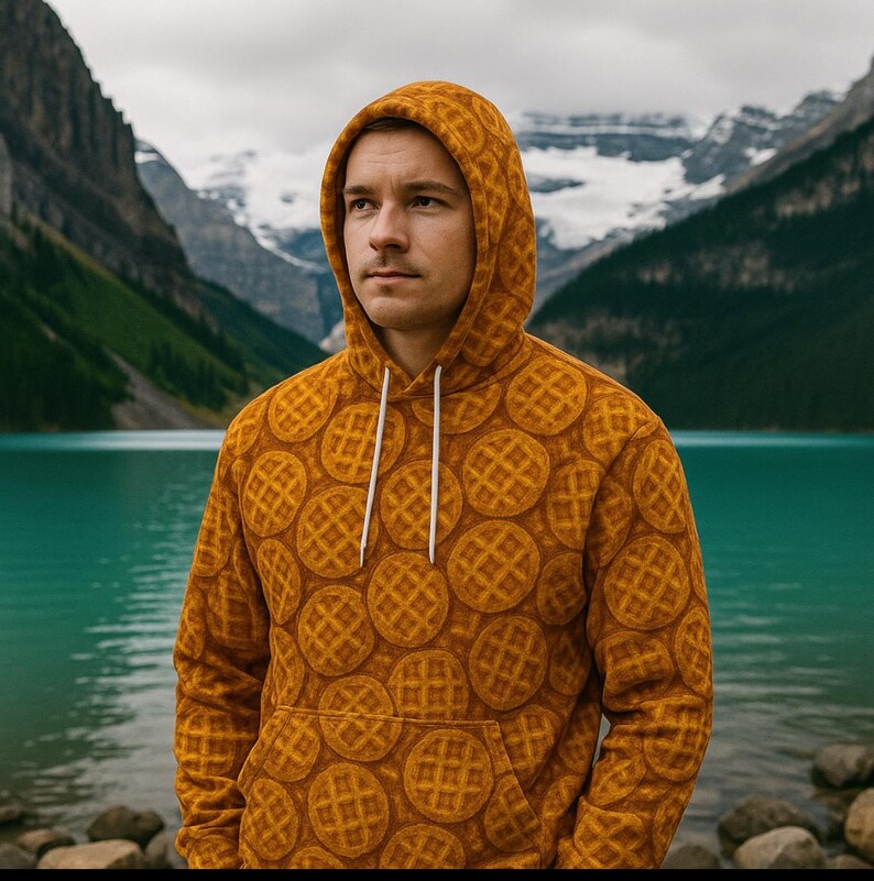 May include: A mustard-colored hoodie with a waffle pattern. The hoodie has a drawstring hood and a front pocket. The waffle design is a repeating pattern of circular waffles. The background features a lake and mountains.