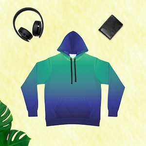 Puede incluir: Una sudadera con capucha degradada en tonos azules y verdes, con un bolsillo delantero y una capucha con cordón. La imagen también incluye auriculares negros y una cartera negra, todo sobre un fondo amarillo claro.
