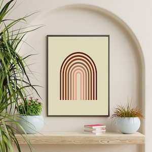 Op de afbeelding: Een ingelijste kunstdruk met een minimalistisch ontwerp van een bruine boog op een crèmekleurige achtergrond. Het kunstwerk wordt tentoongesteld op een houten plank, met planten in blauwe potten en een stapel boeken. De algehele esthetiek is modern en decoratief.