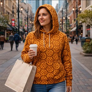 May include: A hooded sweatshirt with a waffle pattern in shades of orange and brown. The sweatshirt has a drawstring hood and a front pocket. The person is holding a white coffee cup and a brown paper shopping bag.