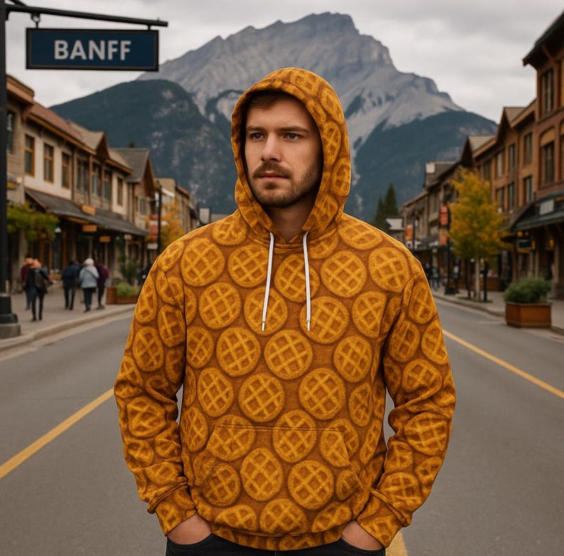 May include: A mustard-yellow hoodie with a repeating waffle pattern. The hoodie has a drawstring and a front pocket. The word "BANFF" is visible on a street sign in the background. The hoodie is worn by a person standing on a street.