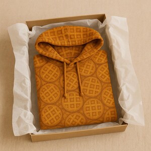 May include: Mustard yellow hoodie with a waffle pattern, presented in a cardboard box with white tissue paper. The hoodie features a drawstring hood and a repeating circular waffle design. The overall aesthetic is warm and inviting.