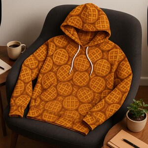 May include: A hooded sweatshirt featuring a repeating pattern of waffles in shades of brown and orange. The hoodie has a front pocket and white drawstrings. The garment is displayed on a dark gray armchair.