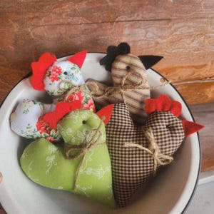 May include: A collection of handmade fabric chicken ornaments in a white enamel bowl. The chickens feature red combs, black or brown eyes, and are made from various patterned fabrics, including floral and checkered designs. The bowl sits on a wooden surface.