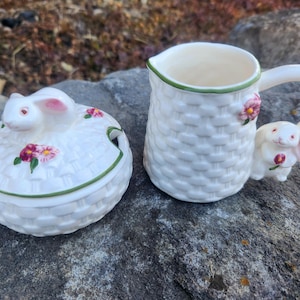May include: White ceramic sugar bowl and creamer set with a woven basket design and bunny rabbit accents. The lid of the sugar bowl features a pink-nosed bunny with pink ears. The creamer has a bunny handle.