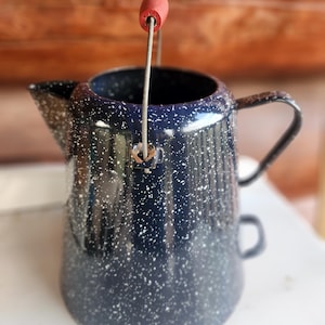 May include: A navy blue enamelware coffee pot with white speckles. It has a black handle and a red wooden handle on the wire bail. The spout is on the left side of the pot.