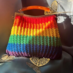 Könnte beinhalten: Handgefertigte, regenbogenfarbene Häkeltasche mit Schildpattgriff und goldfarbenem Metallverschluss. Die Tasche hat ein lebendiges Farbspektrum und ist mit dekorativen Schmetterlingsverzierungen versehen.