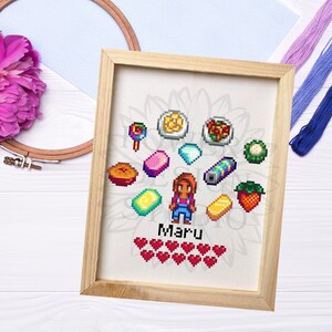 May include: Cross-stitch artwork in a wooden frame featuring a pixelated character named Maru, surrounded by various pixelated food items and objects. The artwork includes a health bar of hearts and is set against a white background.