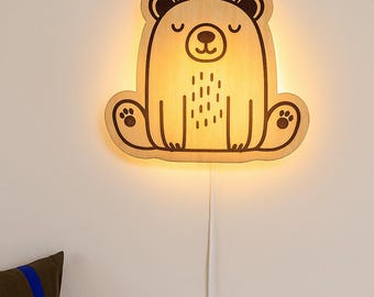 Op de afbeelding: Een houten beer-vormige wandlamp die een warm licht uitstraalt. De beer heeft een eenvoudig, cartoonachtig ontwerp met geëtste details, waaronder gesloten ogen, een neus en pootafdrukken. De lamp is aangesloten op een stopcontact.