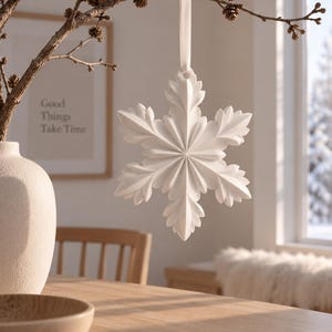 Op de afbeelding: Een matwit sneeuwvlokornament met complexe, gelaagde, bladachtige punten, opgehangen aan een bijpassend wit lint. Het ornament heeft een symmetrisch, stervormig ontwerp met diepe, strak gesneden groeven die vanuit het midden uitstralen.