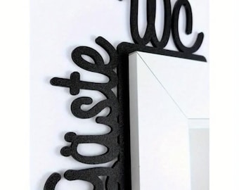 May include: Black metal wall decor spelling out "guest" and "we" in a cursive font. The decor is mounted on a white wall and wraps around the corner of a white frame.