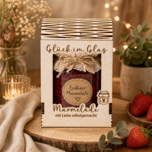 May include: A jar of strawberry marmalade in a wooden box. The jar is sealed with a fabric lid and twine. The box has the words "Glück im Glas" and "Erdbeer Marmelade" printed on it. The box is sitting on a wooden surface.