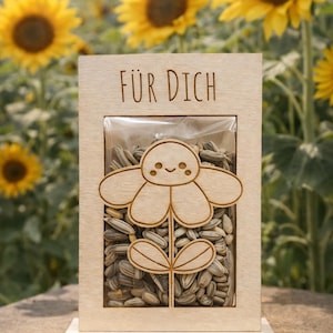Op de afbeelding: Houten doos met een transparant venster gevuld met zonnebloempitten. Een lasergesneden zonnebloemontwerp staat op de voorkant. De woorden "FÜR DICH" zijn bovenaan gegraveerd. De achtergrond toont een zonnebloemveld.
