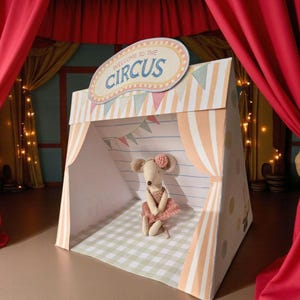 Puede incluir: Un juego de circo con una carpa a rayas y un letrero de "Bienvenido al circo". Dentro, una muñeca de ratón de peluche con tutú rosa sentada en un suelo a cuadros. Cortinas rojas y banderines.