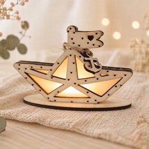May include: A wooden boat-shaped decorative light with a warm glow. The boat has a flag with a heart and anchor detail. The light sits on a round wooden base and is surrounded by soft, neutral-toned decor.