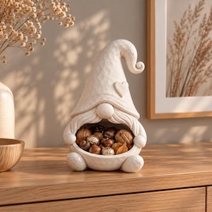 Könnte beinhalten: Eine cremefarbene Zwergenfigur mit geschnitzter Holzoptik, die einen hohen spitzen Hut mit einem kleinen Herzdetail und einen breiten Bart aufweist. Der Zwerg hat eine hohle Mitte, die mit verschiedenen braunen Nüssen, Eicheln und kleinen Pflanzenteilen gefüllt ist.