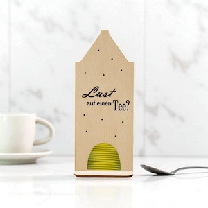 May include: Wooden tea bag holder with a house-shaped design. The holder has the text "Lust auf einen Tee?" and contains yellow tea bags. A white teacup and saucer are visible in the background, along with a spoon.