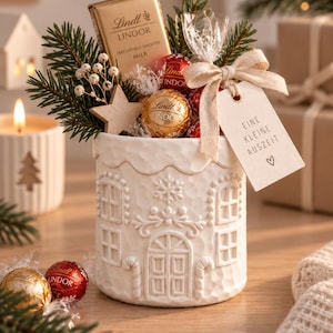 Könnte beinhalten: Ein weißer Keramikbehälter in Hausform mit geprägten Fenstern und Türen, gefüllt mit Lindt-Schokoladentafeln und -kugeln, Tannenzweigen und einem Holzstern. Ein beiges Band hält ein Etikett mit der Aufschrift EINE KLEINE AUSZEIT und einem Herz.